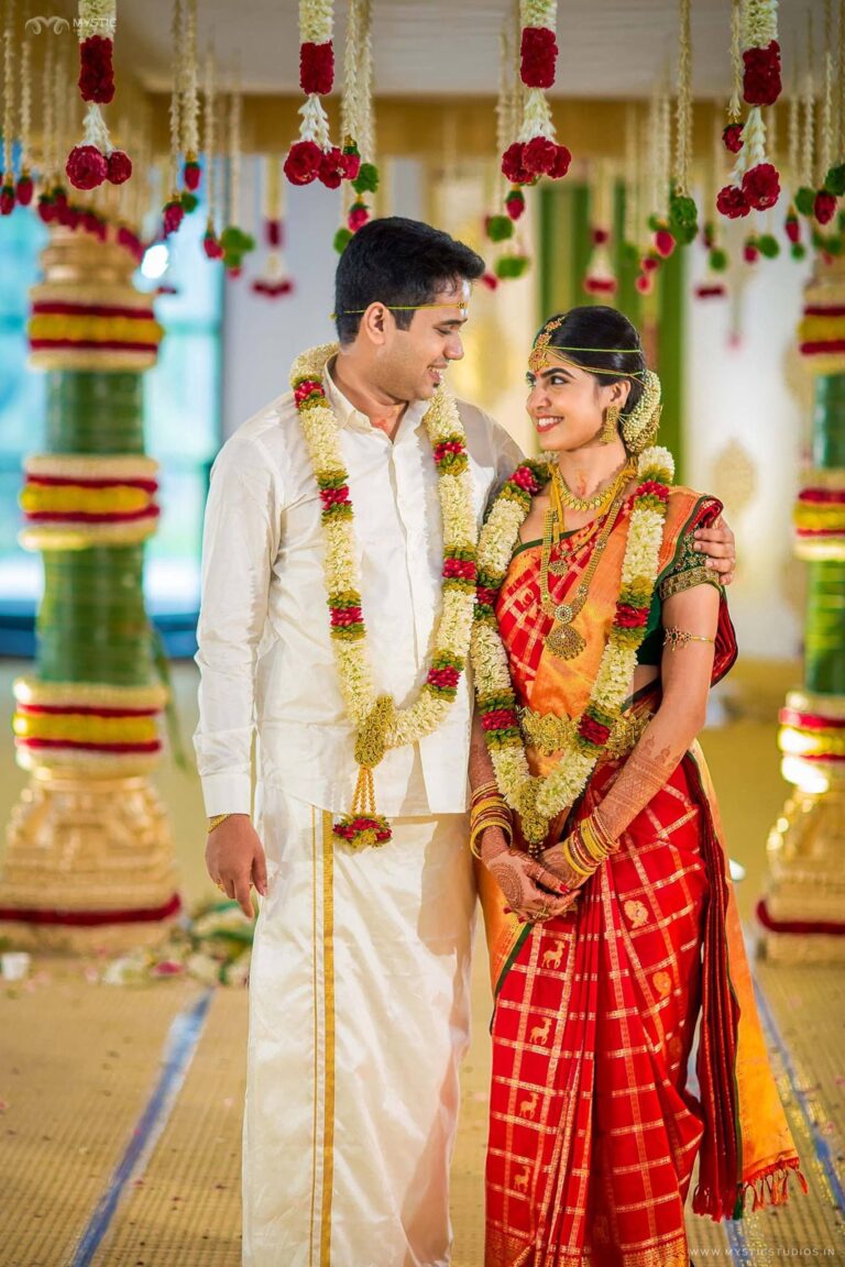 Happy Telugu wedding couple dressed in traditional white and red silk attire, exchanging smiles during ceremony with beautiful floral decorations — best wedding photographers in Hyderabad.